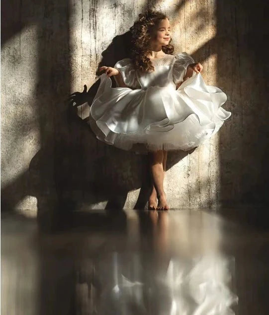 Child in puffy white flower girl dress standing in sunlight, Babelio brand, indoor setting